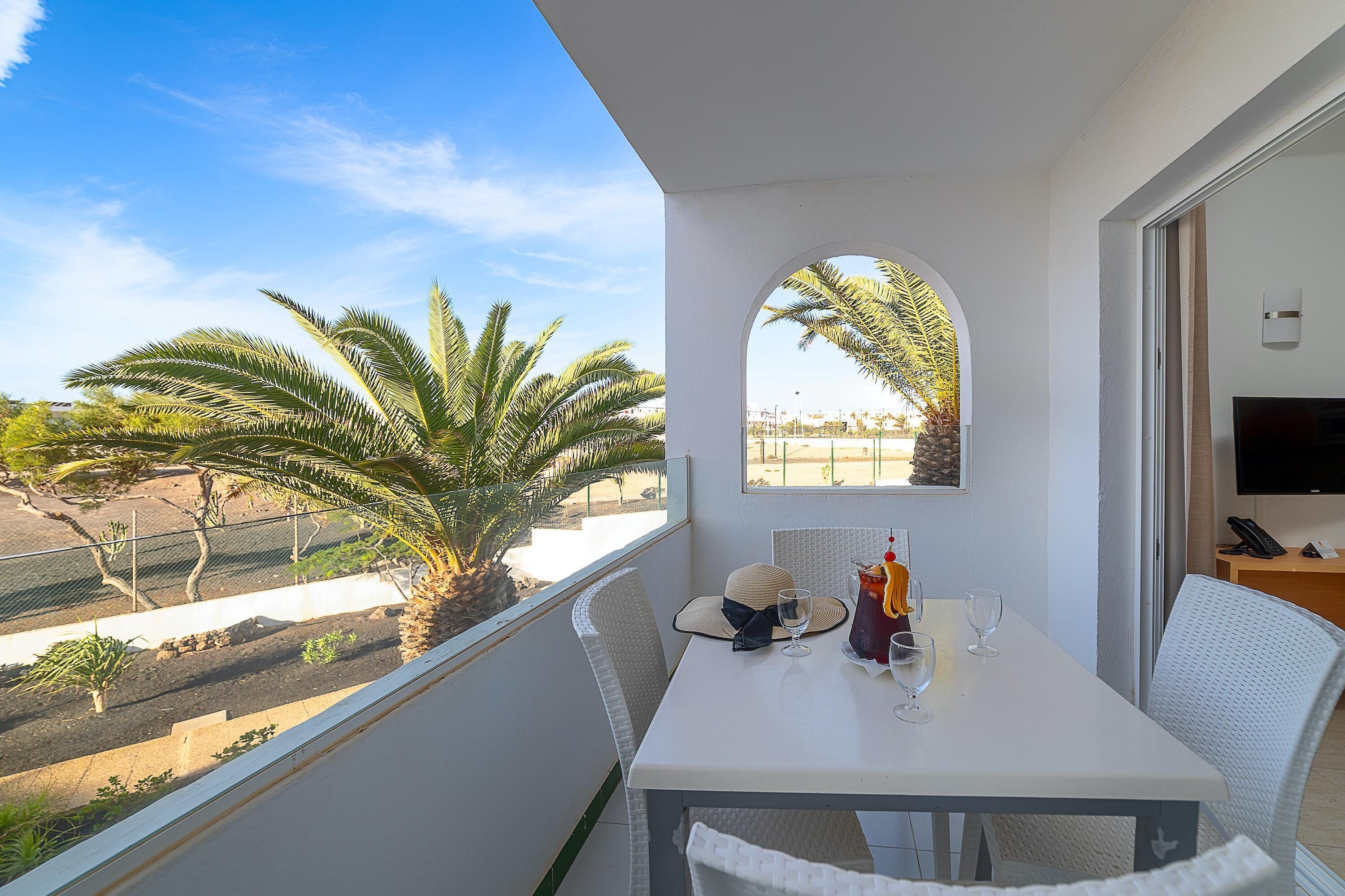 A balcony with a white table, two chairs, a TV, and a view of palm trees and a beach.