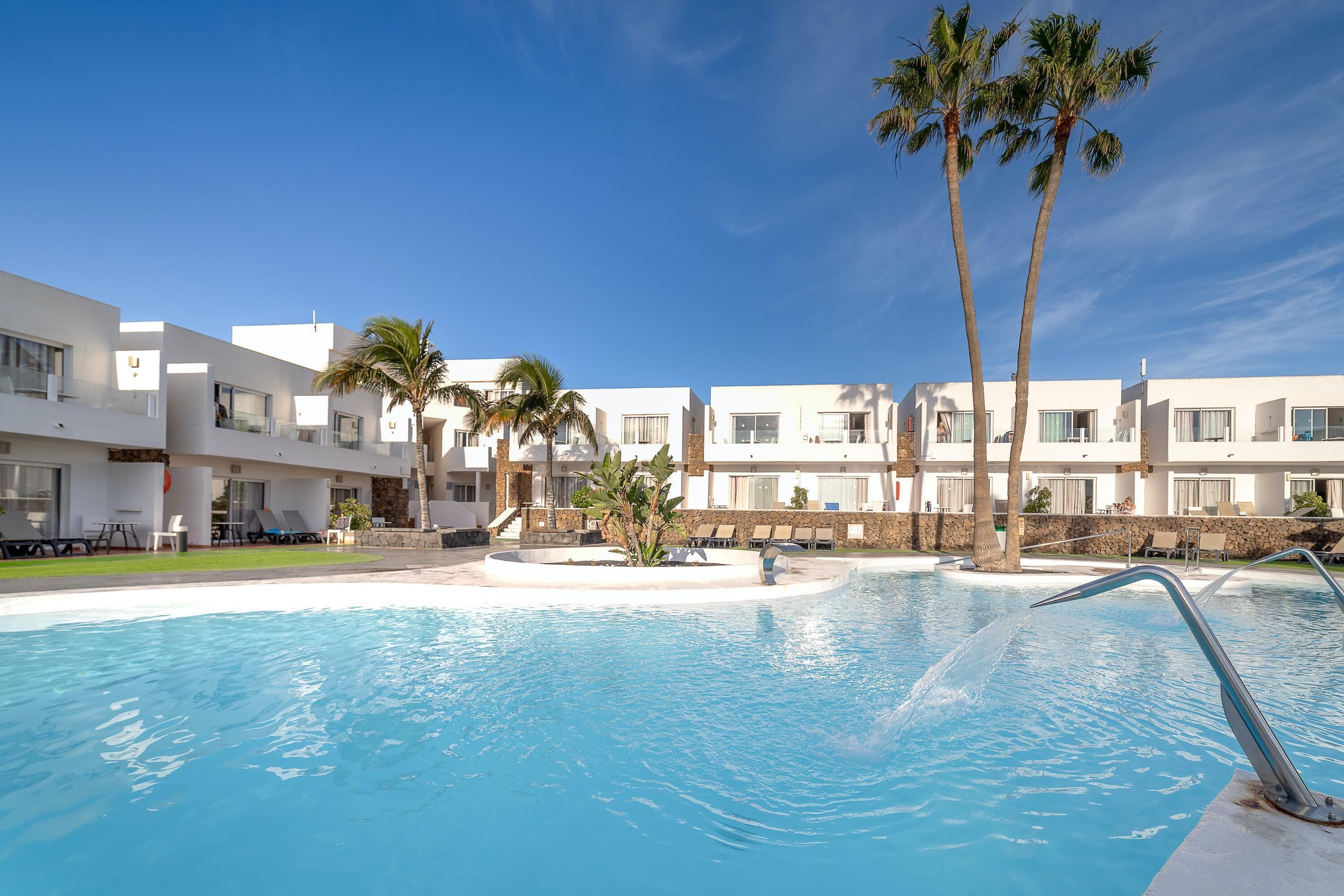 Un hotel moderno con piscina, palmeras y un cielo azul despejado.