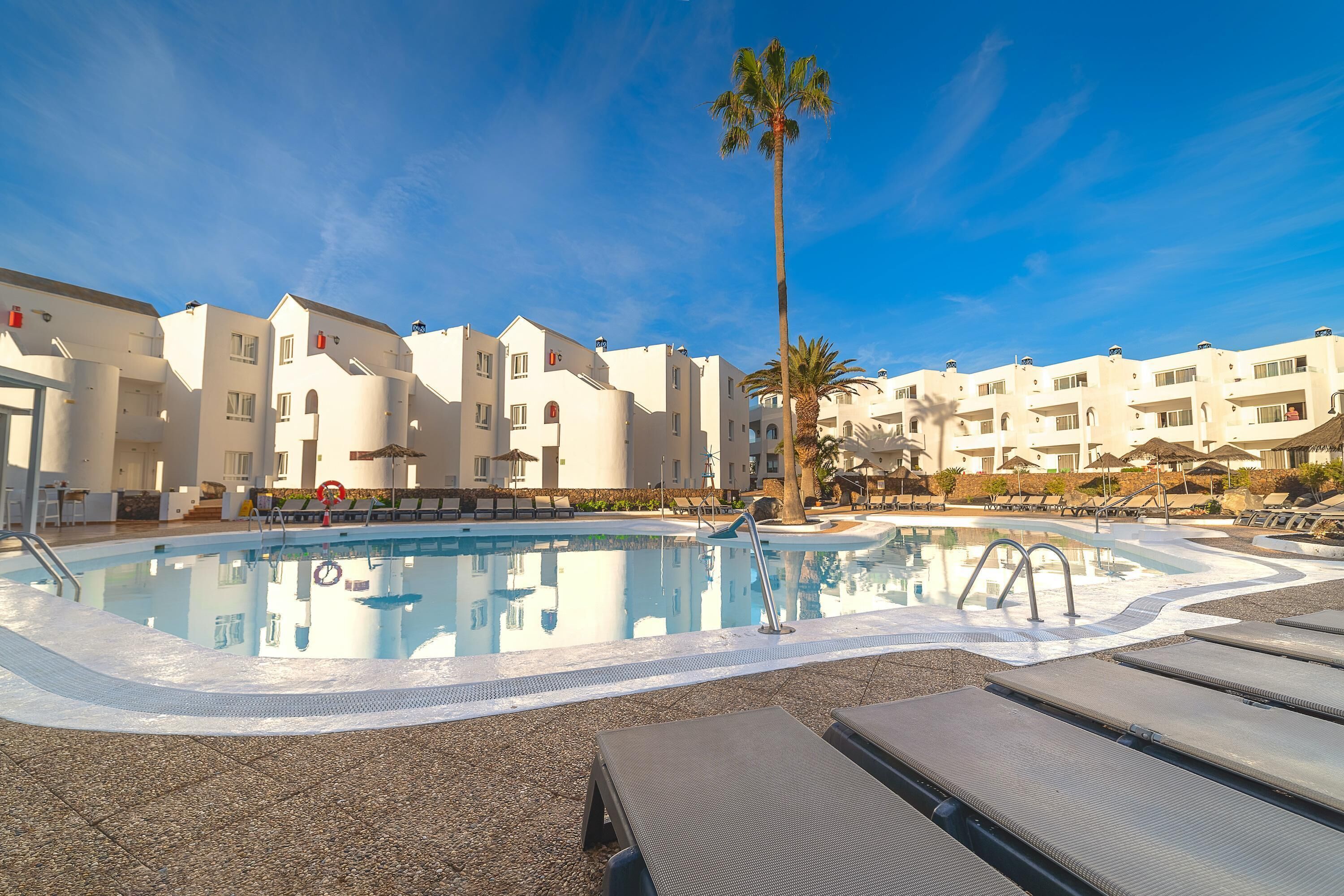 A swimming pool with lounge chairs, a palm tree, and white buildings in the background.