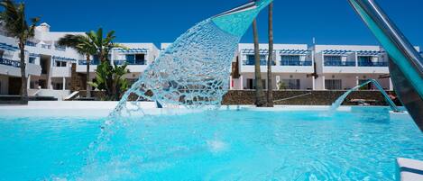 A pool with clear blue water, a metal structure, and a building with balconies and palm trees in the background.