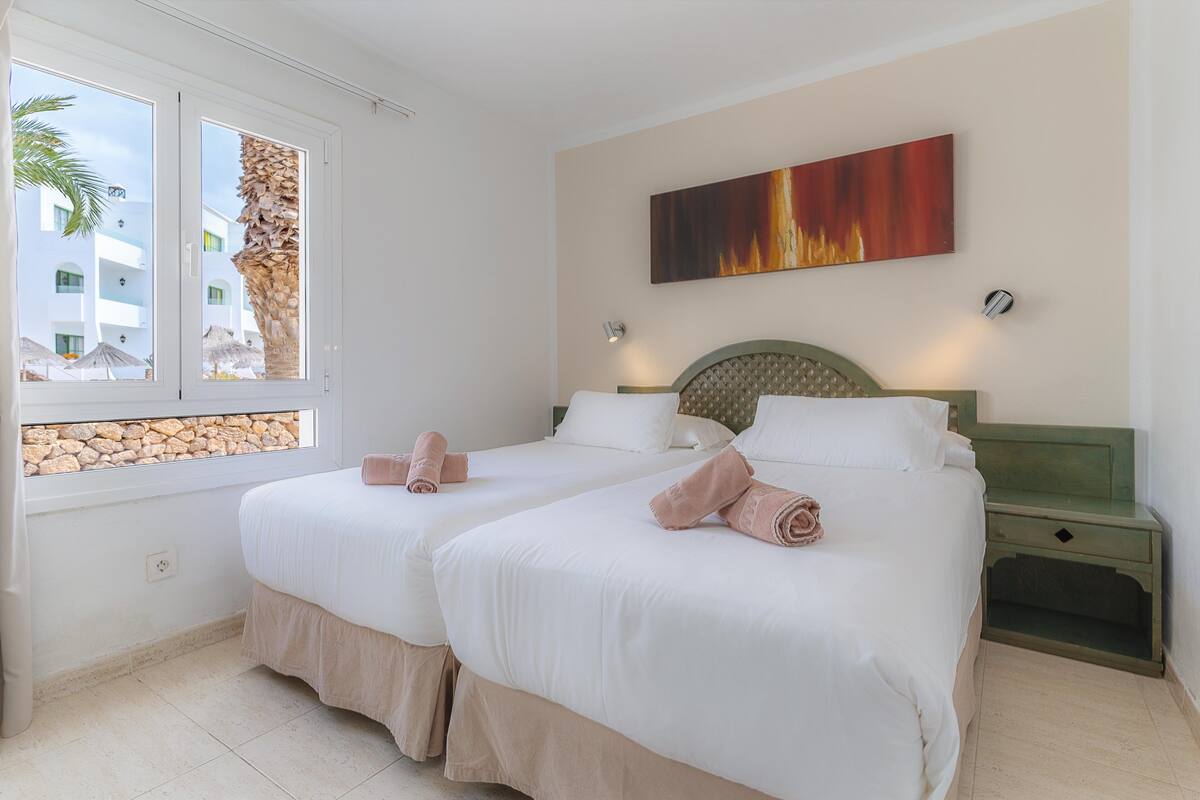 A hotel room with two beds, a stone wall, a window with a view of buildings and palm trees, and a painting on the wall.