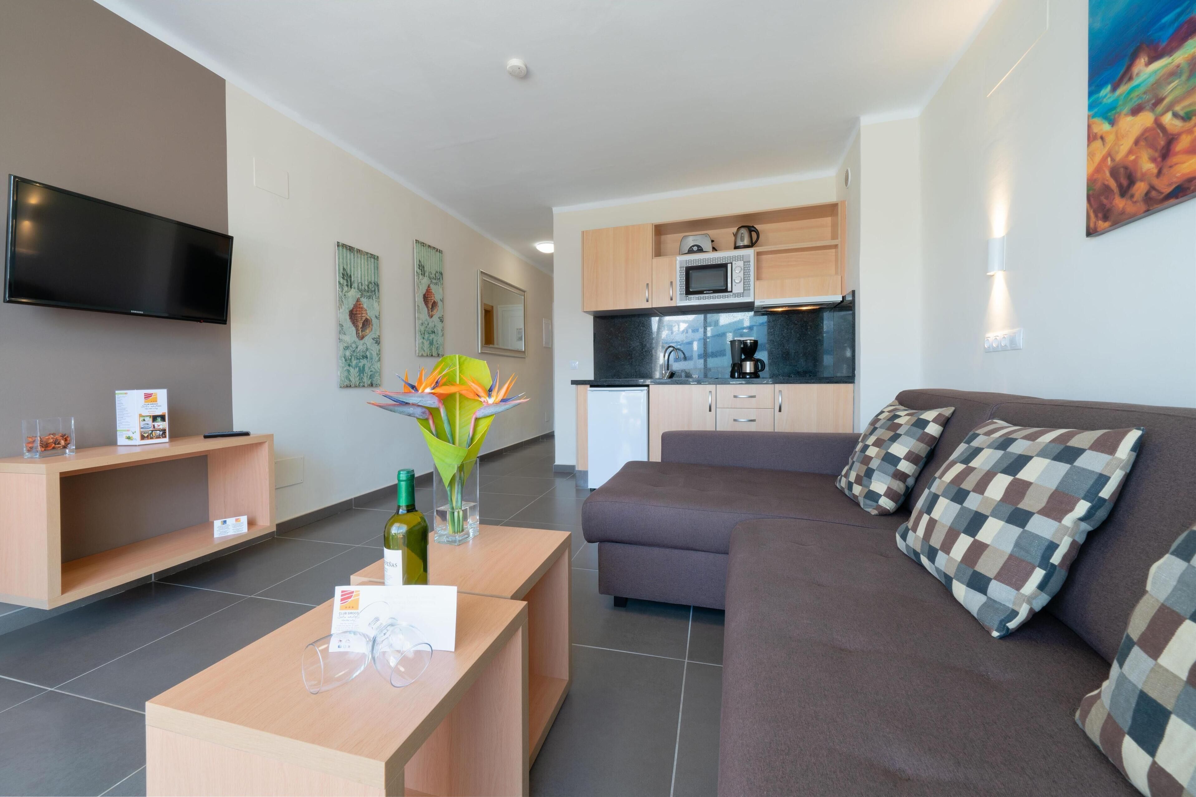 Una sala de estar moderna con un sofá, una mesa de centro y un televisor. Cuenta con una zona de cocina con armarios y un microondas.