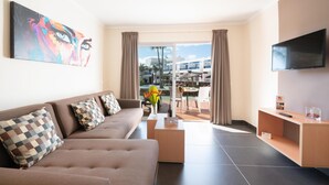 A modern living room with a sofa, a coffee table, and a flat-screen TV. There is a balcony with a view of buildings and palm trees. - Hotel Siroco - Adults Only (Teguise)