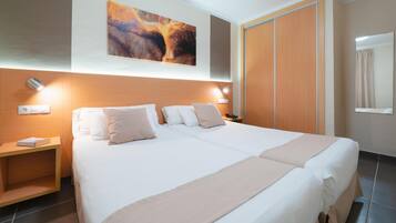 Un lit double avec une couvre-lit et des oreillers de couleur beige, une table de chevet avec un livre, une lampe fixée au mur et une armoire en bois.