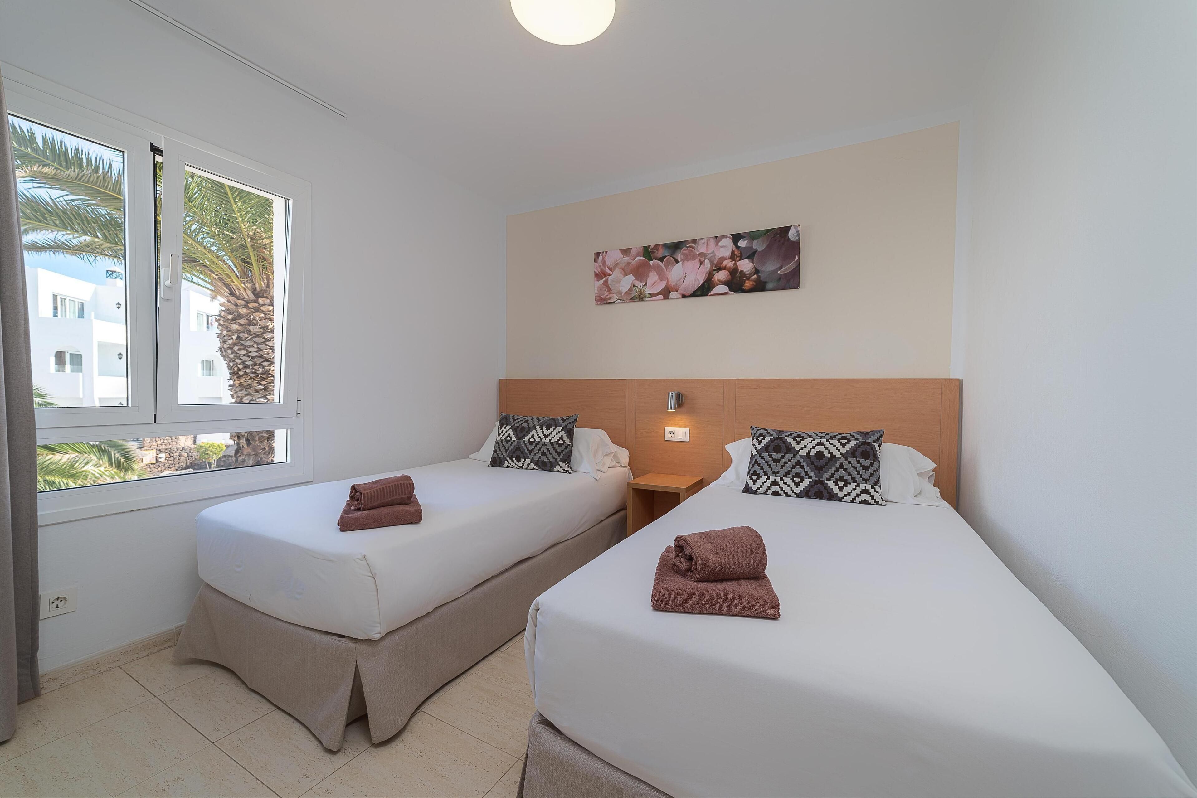 A hotel room with two single beds, a window with a view of palm trees, a painting on the wall, and a nightstand with a lamp.
