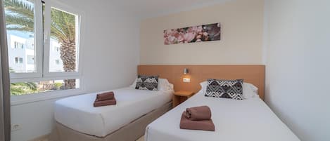 A hotel room with two single beds, a window with a view of palm trees, a painting on the wall, and a nightstand with a lamp.