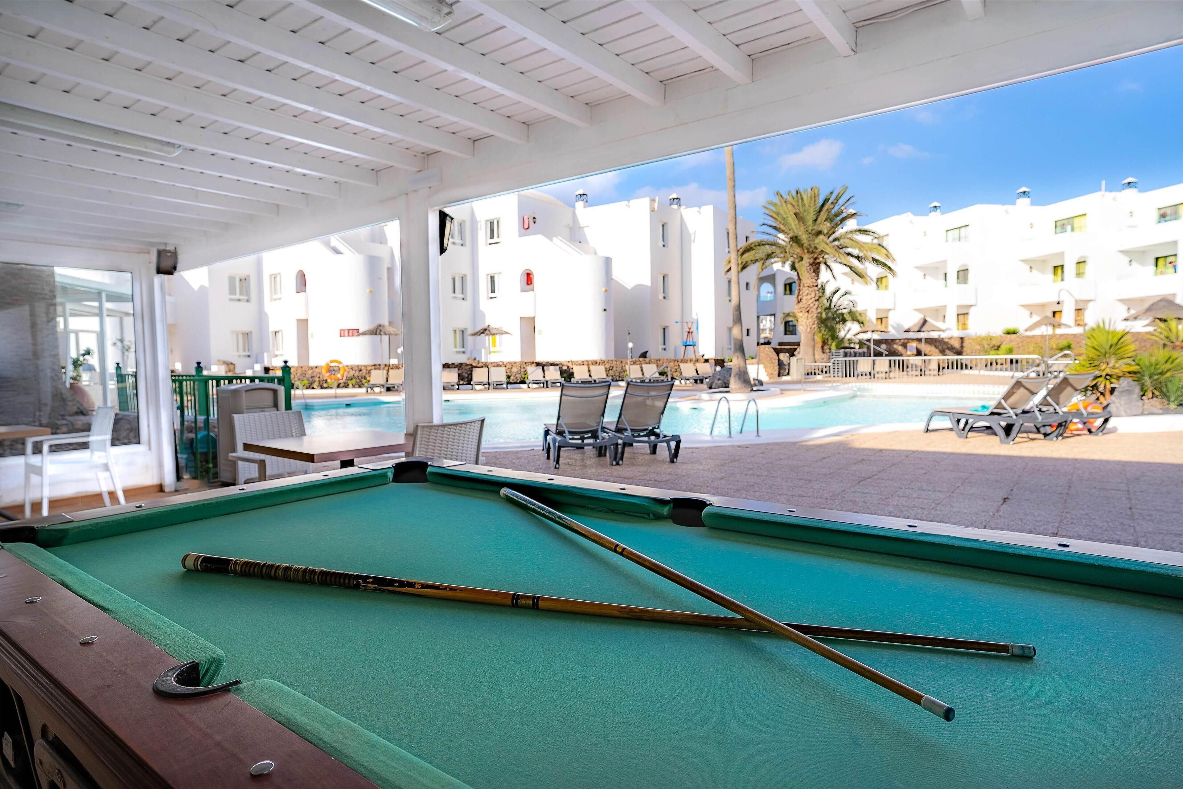 A pool table with pool cues, a pool, and lounge chairs in a covered outdoor area with a view of white buildings and palm trees.
