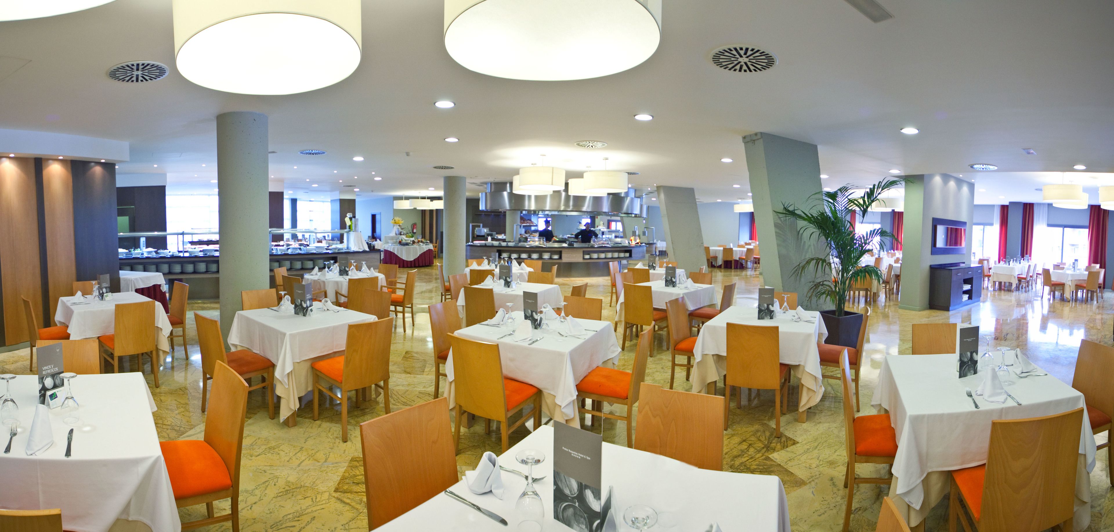 A spacious dining area with multiple tables and chairs, a central kitchen area, and large windows allowing natural light.