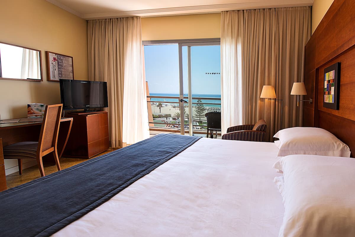 Une chambre d’hôtel avec un grand lit, un bureau avec une télévision, une chaise et une vue sur la plage.