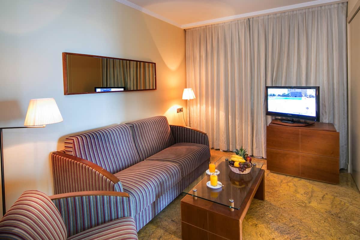 Une chambre d’hôtel avec un canapé à rayures, une table basse en verre sur laquelle sont disposés des fruits, un téléviseur à écran plat et une lampe.