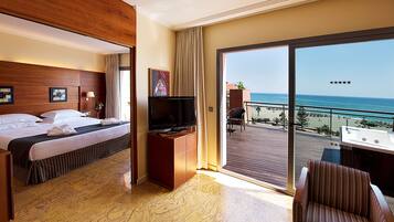 Habitación de hotel con cama, televisor, balcón con vistas al mar y zona de estar.