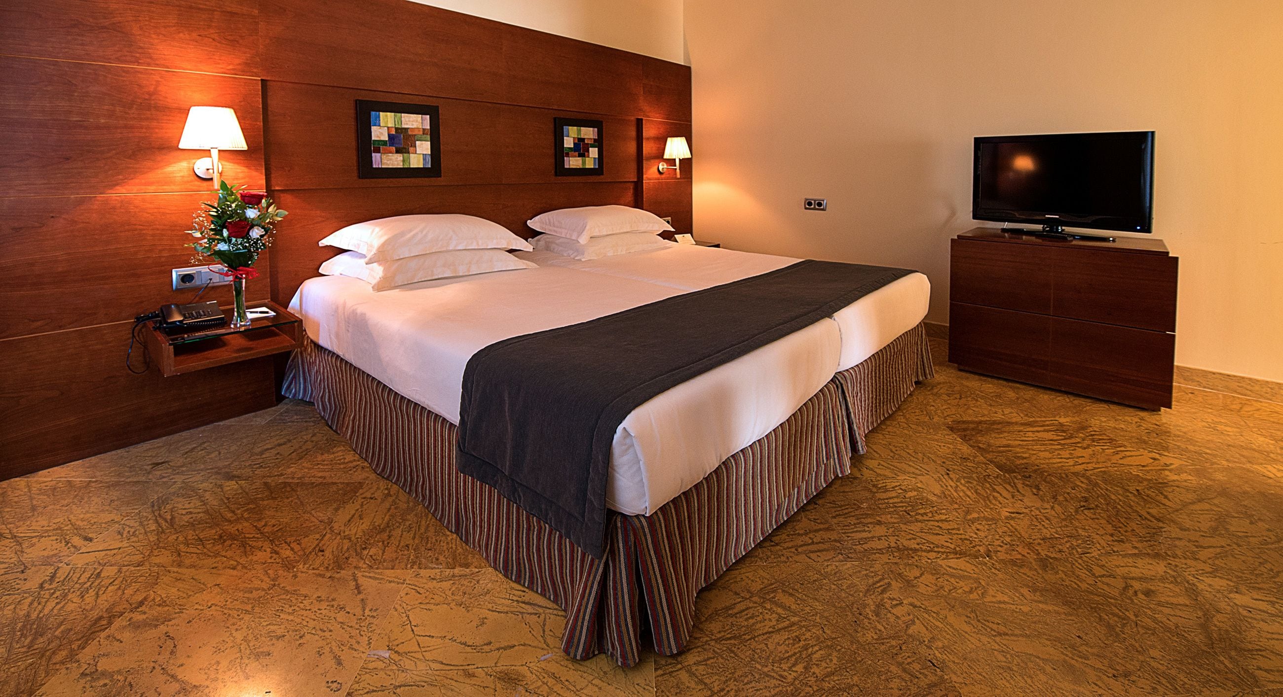 A hotel room with a large bed, a TV, a nightstand with a lamp, and a floral arrangement.