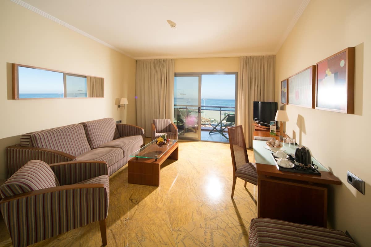 Une chambre d’hôtel avec un canapé, des fauteuils, une table basse, une télévision et un balcon donnant sur la mer.
