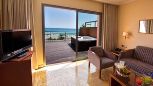Ein Hotelzimmer mit Fernseher, einer Sitzecke, einem Couchtisch mit Getränken sowie einem Balkon mit Blick auf Strand und Meer.