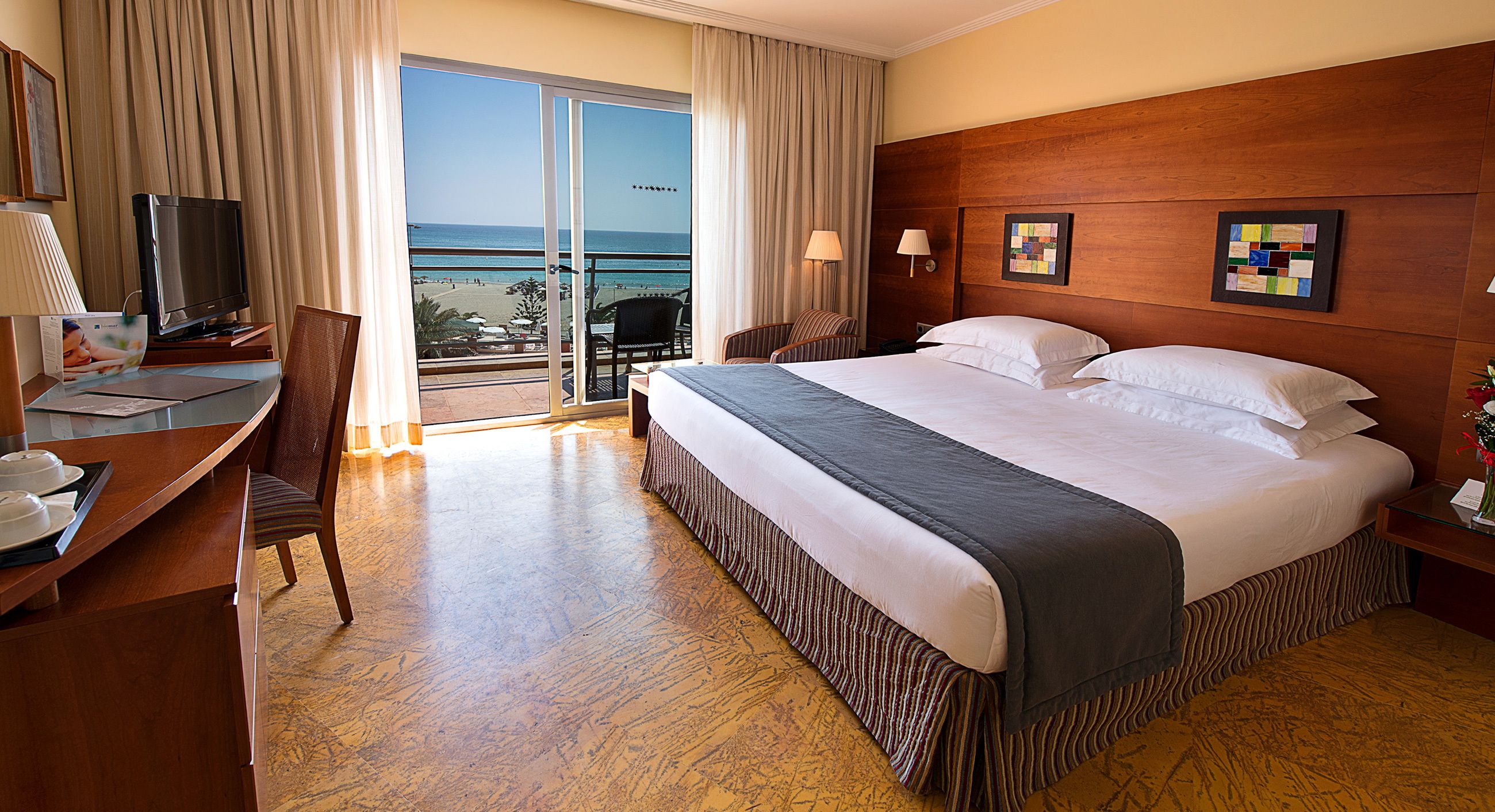 A hotel room with a large bed, a desk with a television, a chair, and a view of the beach through a sliding door.