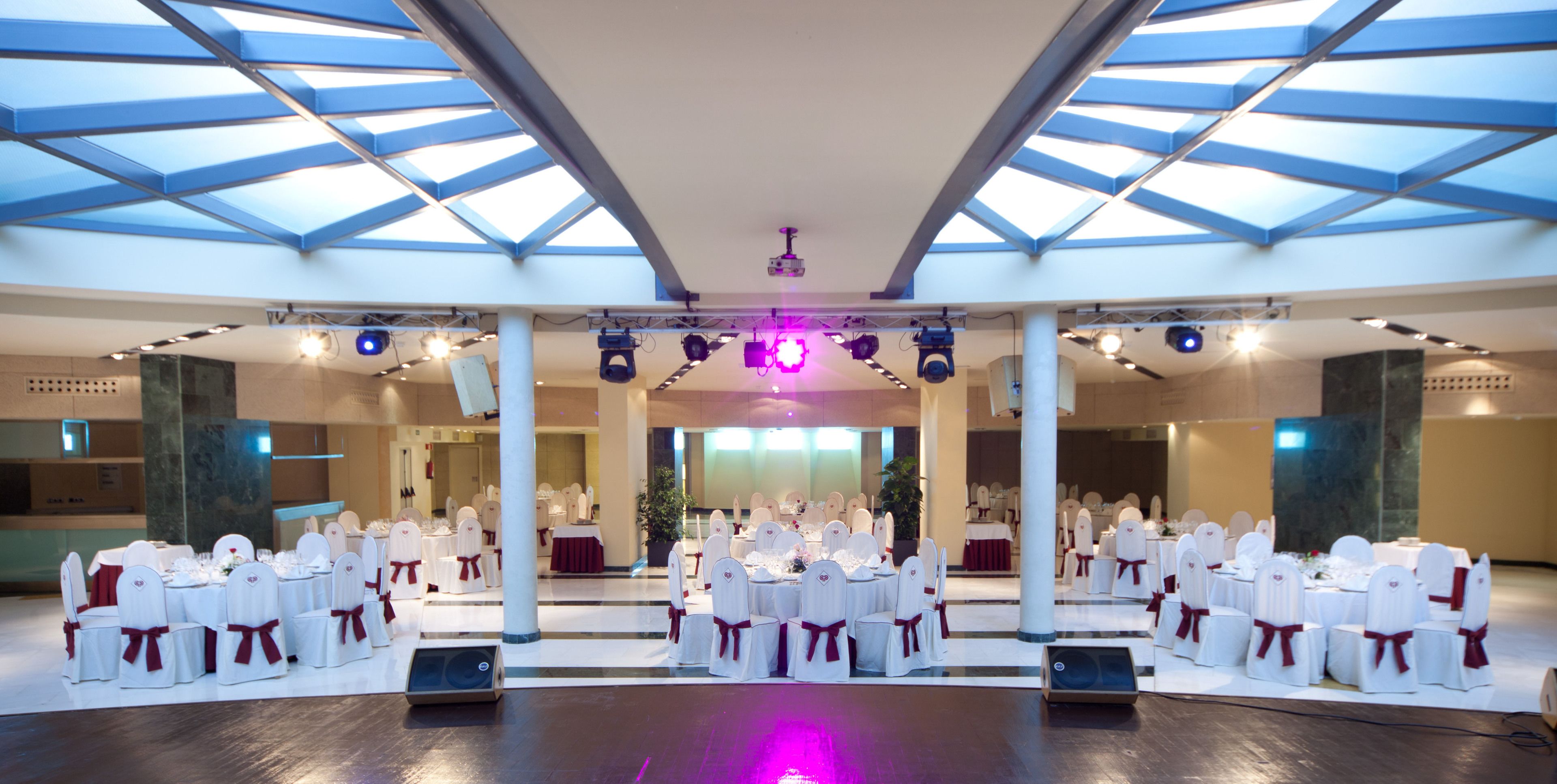 A spacious hall with a high ceiling and skylights, arranged with round tables and white chairs with red sashes, suitable for events.