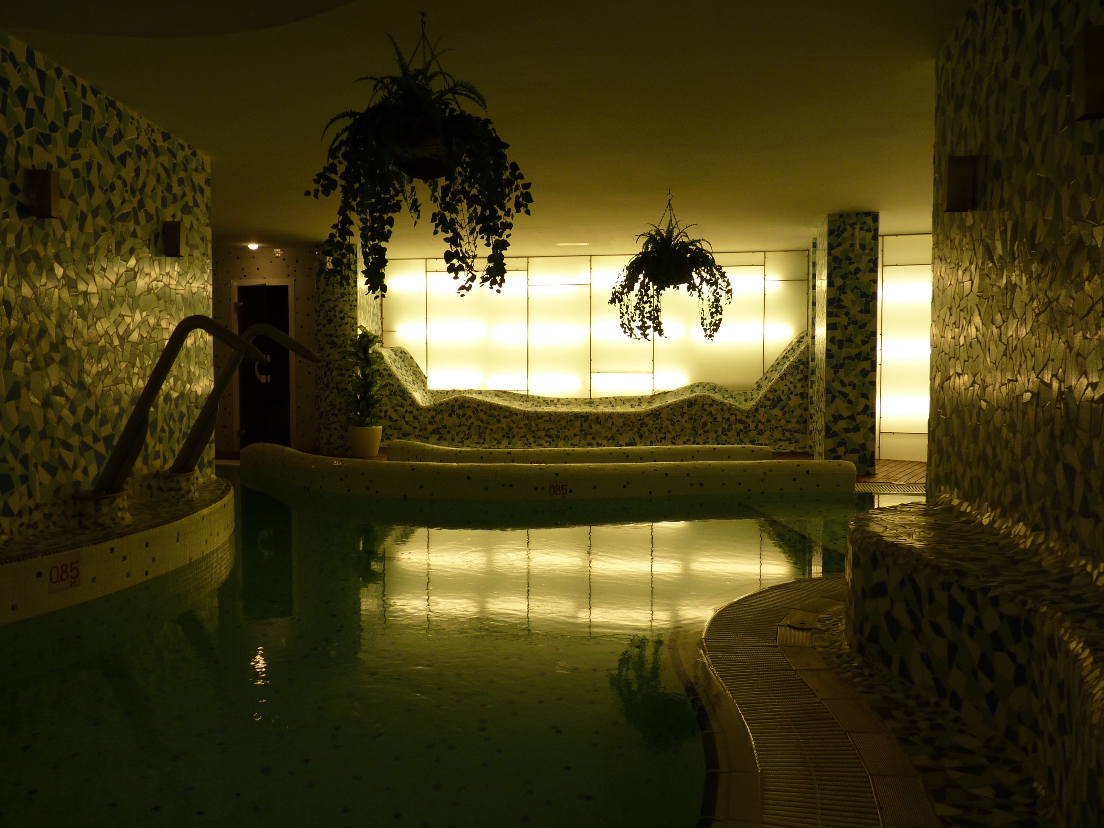 A dimly lit indoor swimming pool with mosaic walls and hanging plants.