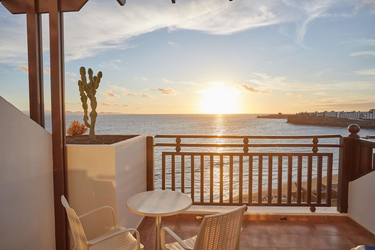 A balcony with a view of the sunset over the sea, a cactus, and wicker furniture.