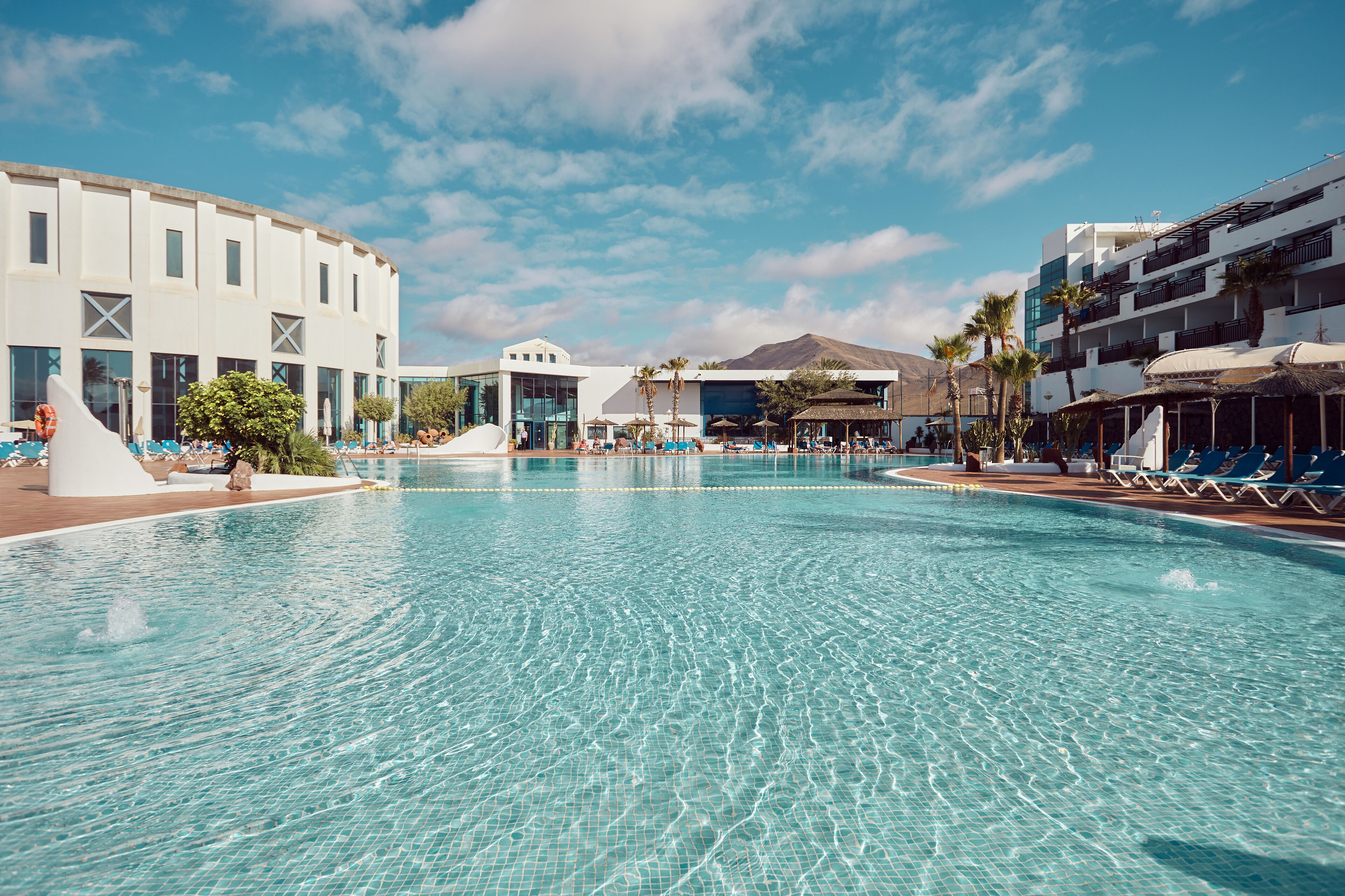 A large swimming pool with clear blue water, surrounded by lounge chairs and palm trees. There are modern buildings with balconies and a mountain in the background.
