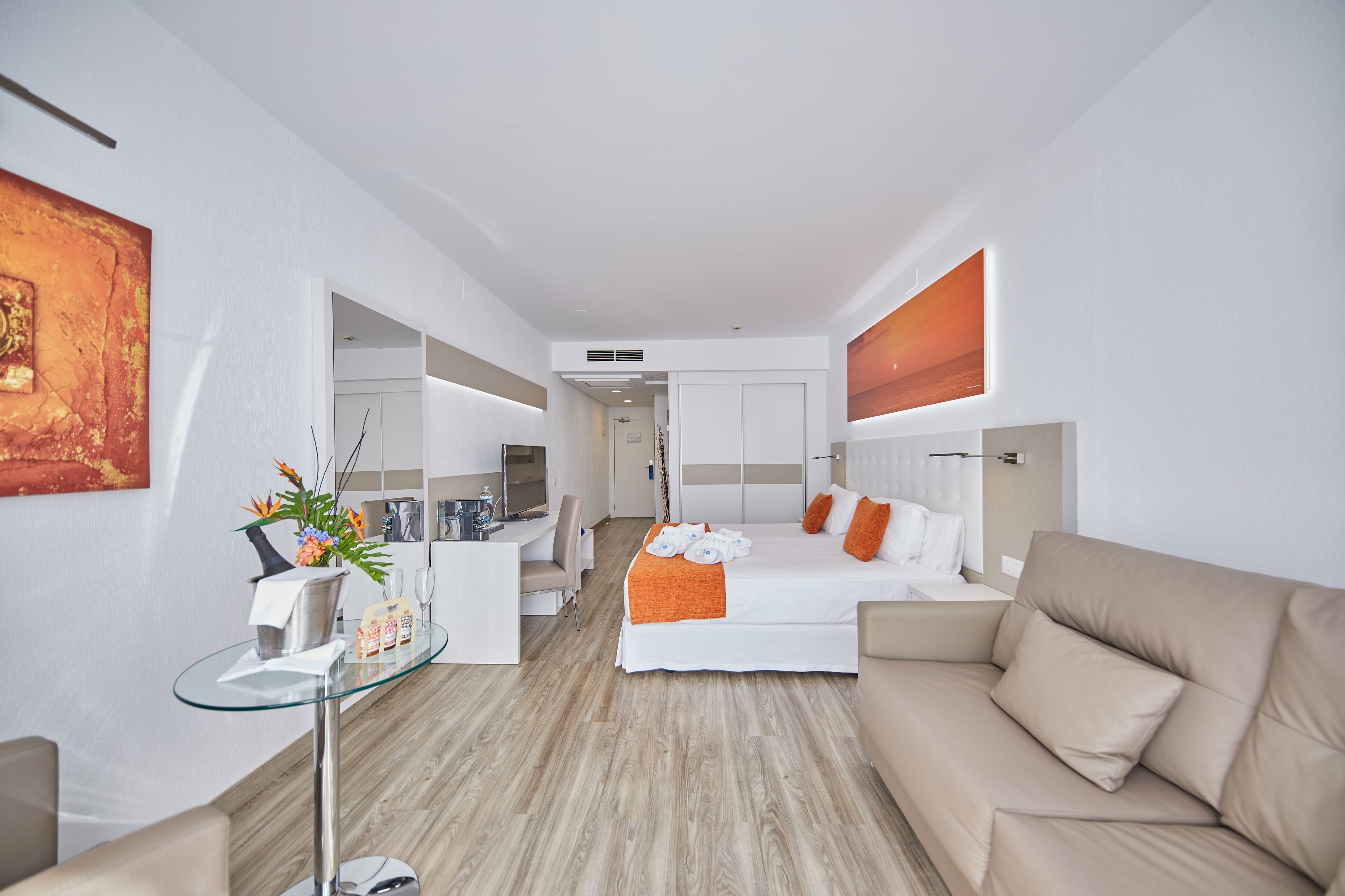 Habitació d'hotel moderna amb un llit, un sofà, una taula de vidre amb un ram de flors i un escriptori amb un ordinador.