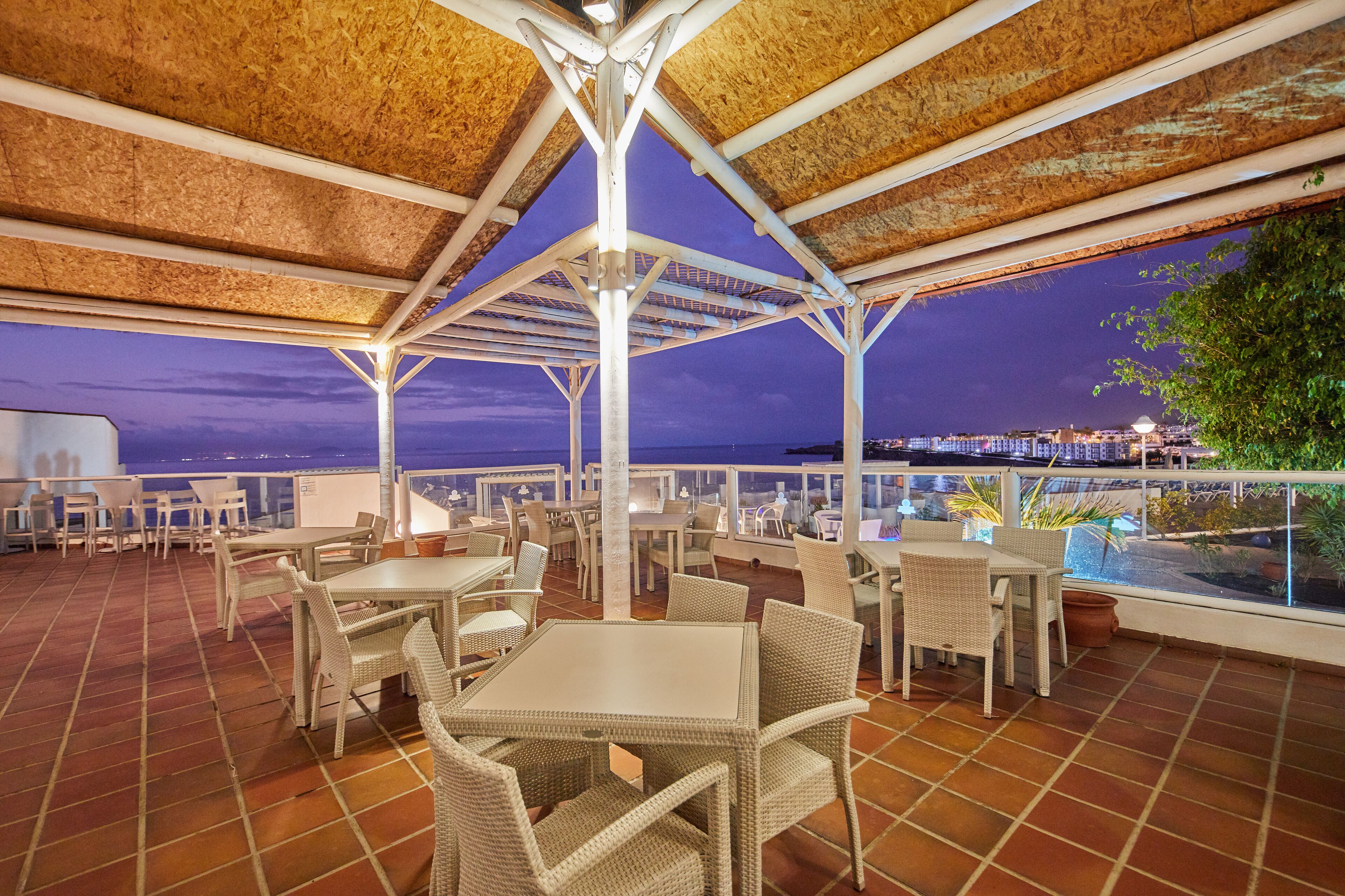 An outdoor terrace with tables and chairs, a tiled floor, and a view of the city at dusk.