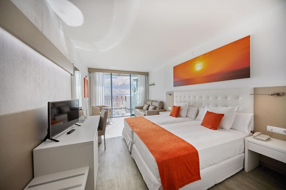 A hotel room with a large bed, a desk with a TV, a chair, a balcony with a view, and a painting of a sunset.