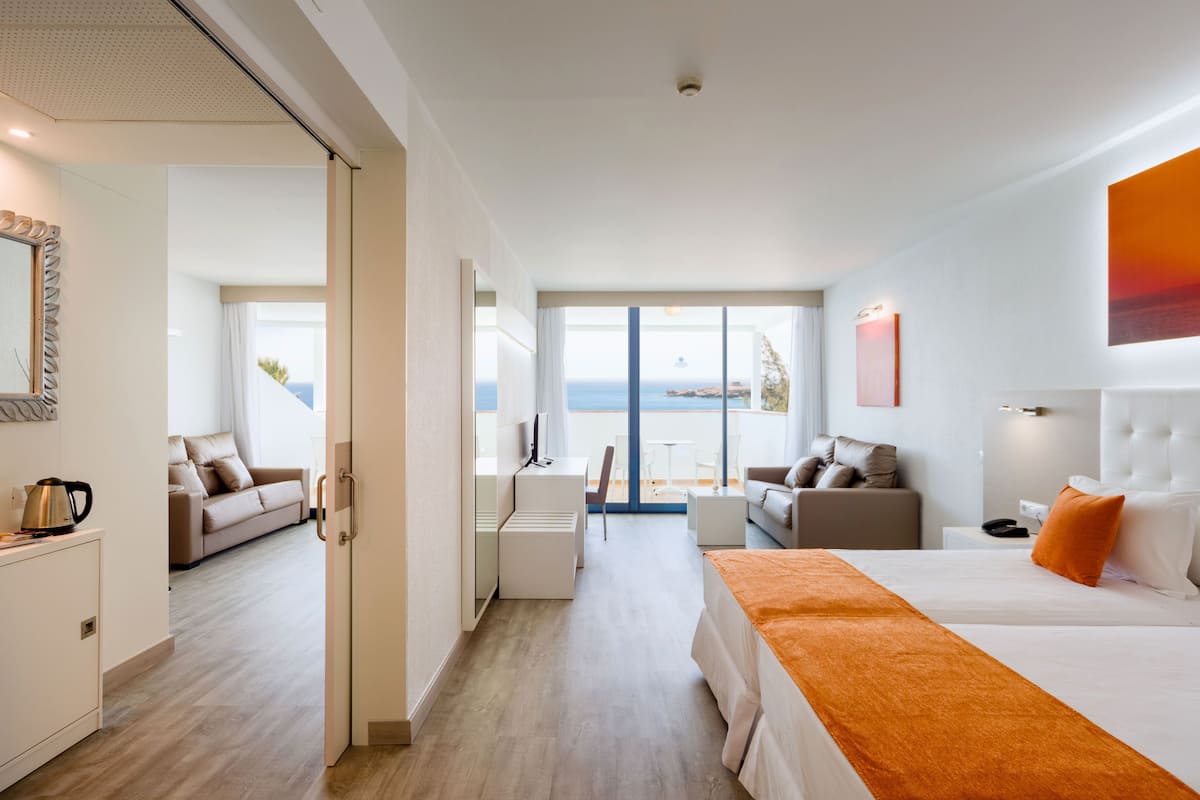 A modern hotel room with a large bed, a sofa, a desk, and a view of the sea.