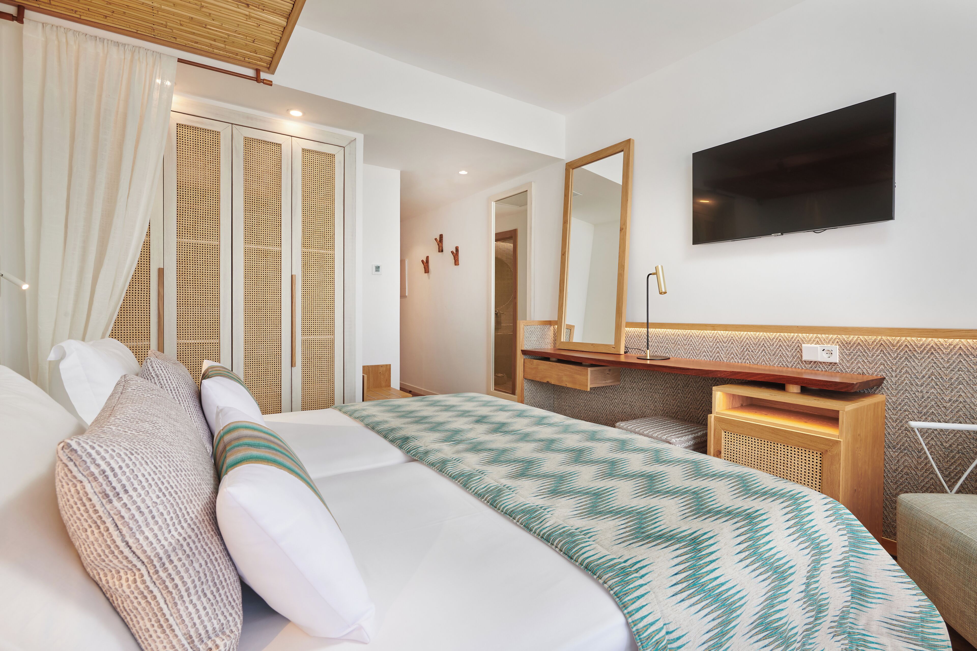 Quarto de hotel com cama, televisão fixada na parede, mesa de cabeceira de madeira e espelho sobre a mesa.