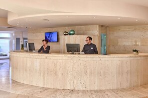 Een receptiebalie met twee medewerkers, een flatscreen televisie en decoratieve vazen.