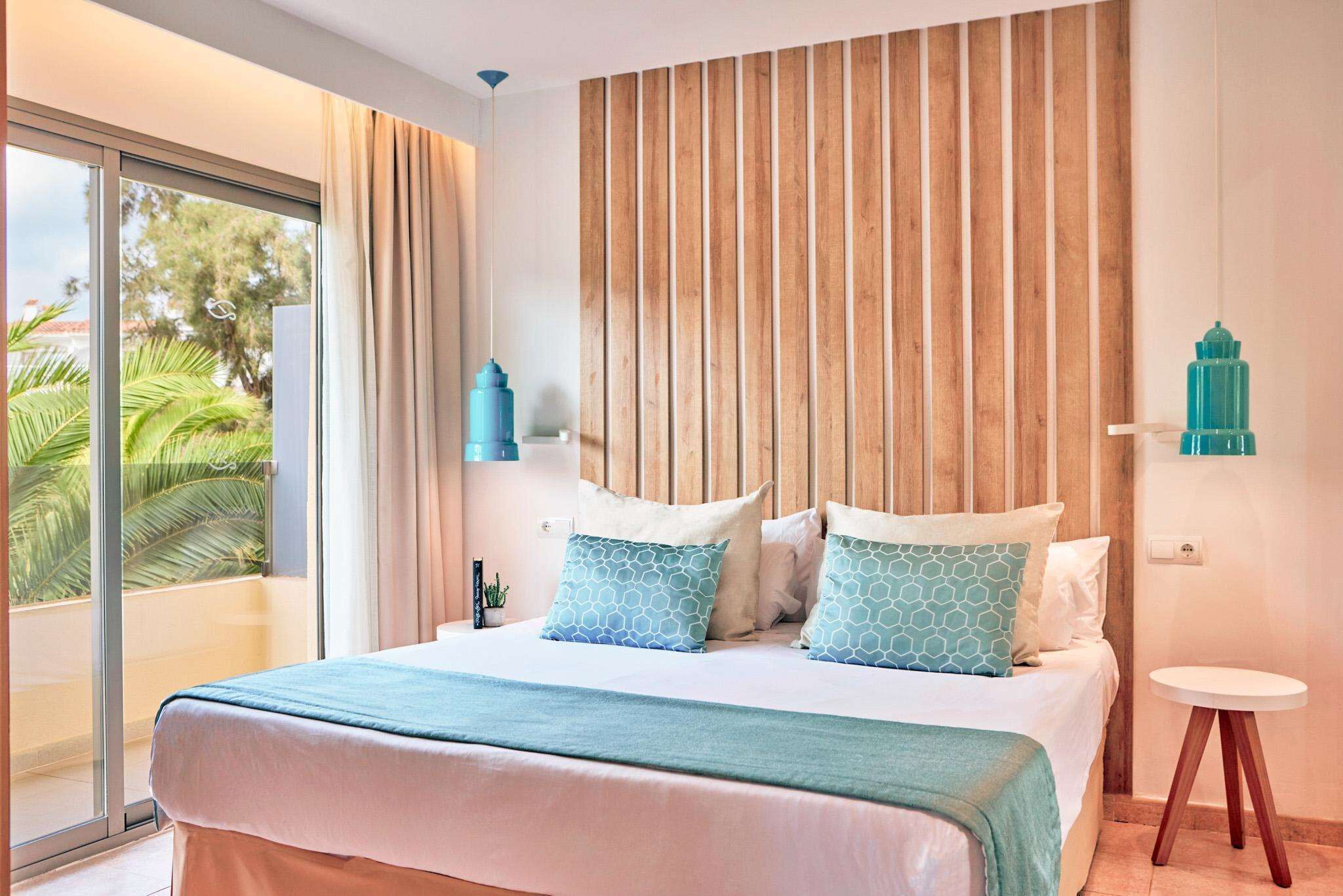 Un dormitorio con una cama grande, cabecera de madera, una mesita redonda y vista a palmeras a través de una puerta corrediza de vidrio.