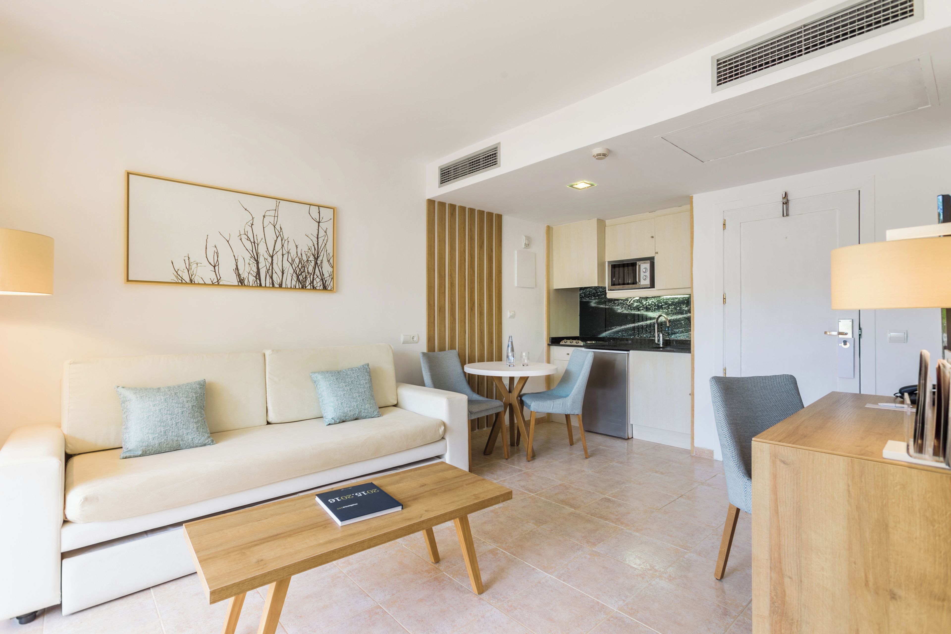 A modern living room with a sofa, coffee table, and chairs. There is a kitchen area with a microwave and cabinets.