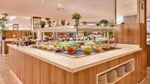 Zona de buffet con diferentes opciones de comida, mostrador de madera y plantas en macetas.