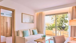 A modern living room with a sofa, a coffee table, and a dining area. There is a large window with curtains, a painting on the wall, and a view of the outdoors. - Zafiro Palmanova (Calvia)