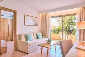 A modern living room with a sofa, a coffee table, and a dining area. There is a large window with curtains, a painting on the wall, and a view of the outdoors. - Zafiro Palmanova (Calvia)