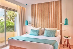 A bedroom with a large bed, a wooden headboard, a view of greenery outside, and a small side table with books. - Zafiro Palmanova (Calvia)