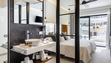 Ett modernt hotellrum med en stor spegel, ett vitt handfat, en träinredd sminkbord, en säng med vita sängkläder och utsikt över hotellets inre.