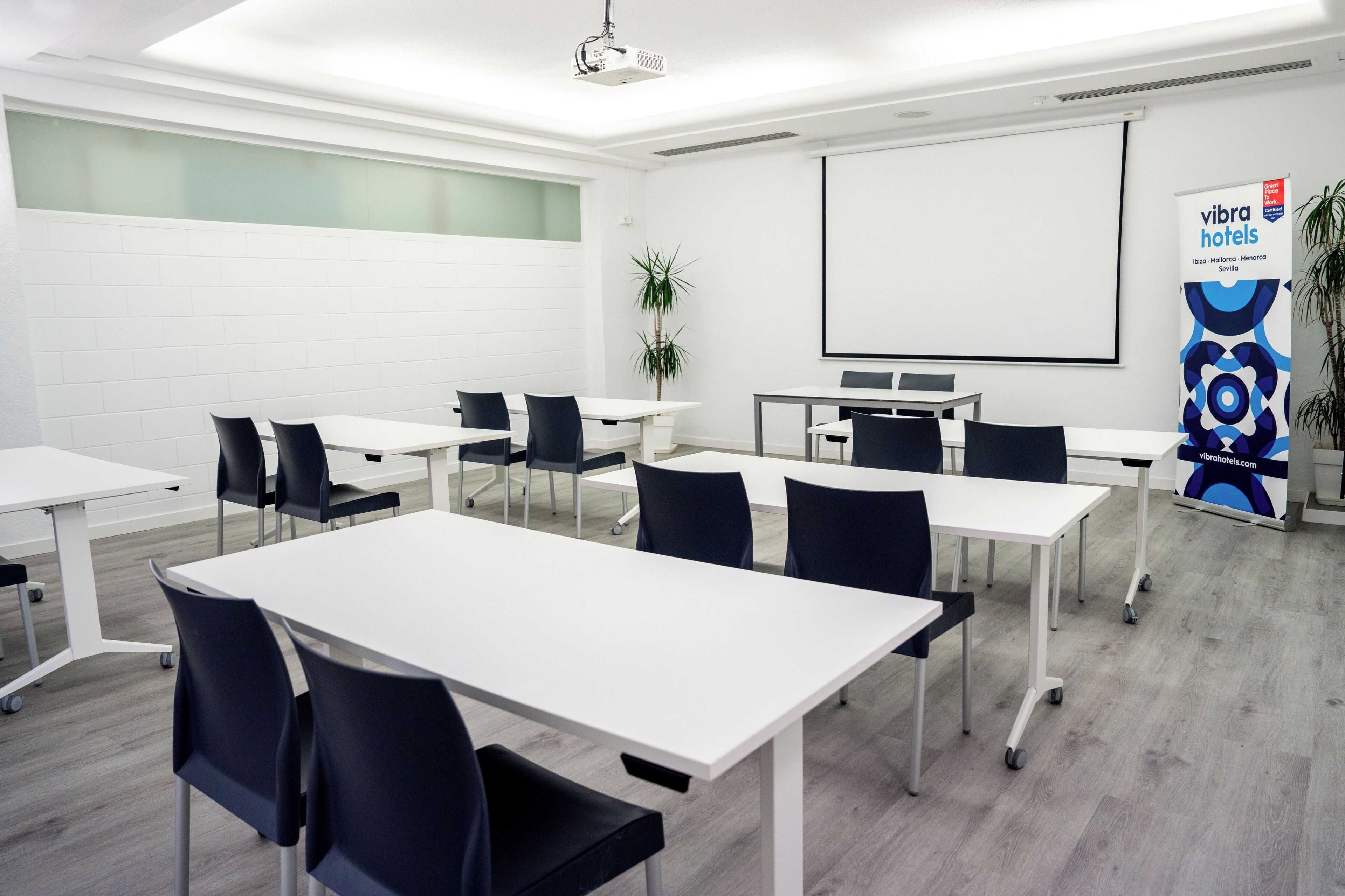 A conference room with white tables, black chairs, a projector, a screen, and a banner for VIBRA HOTELS.