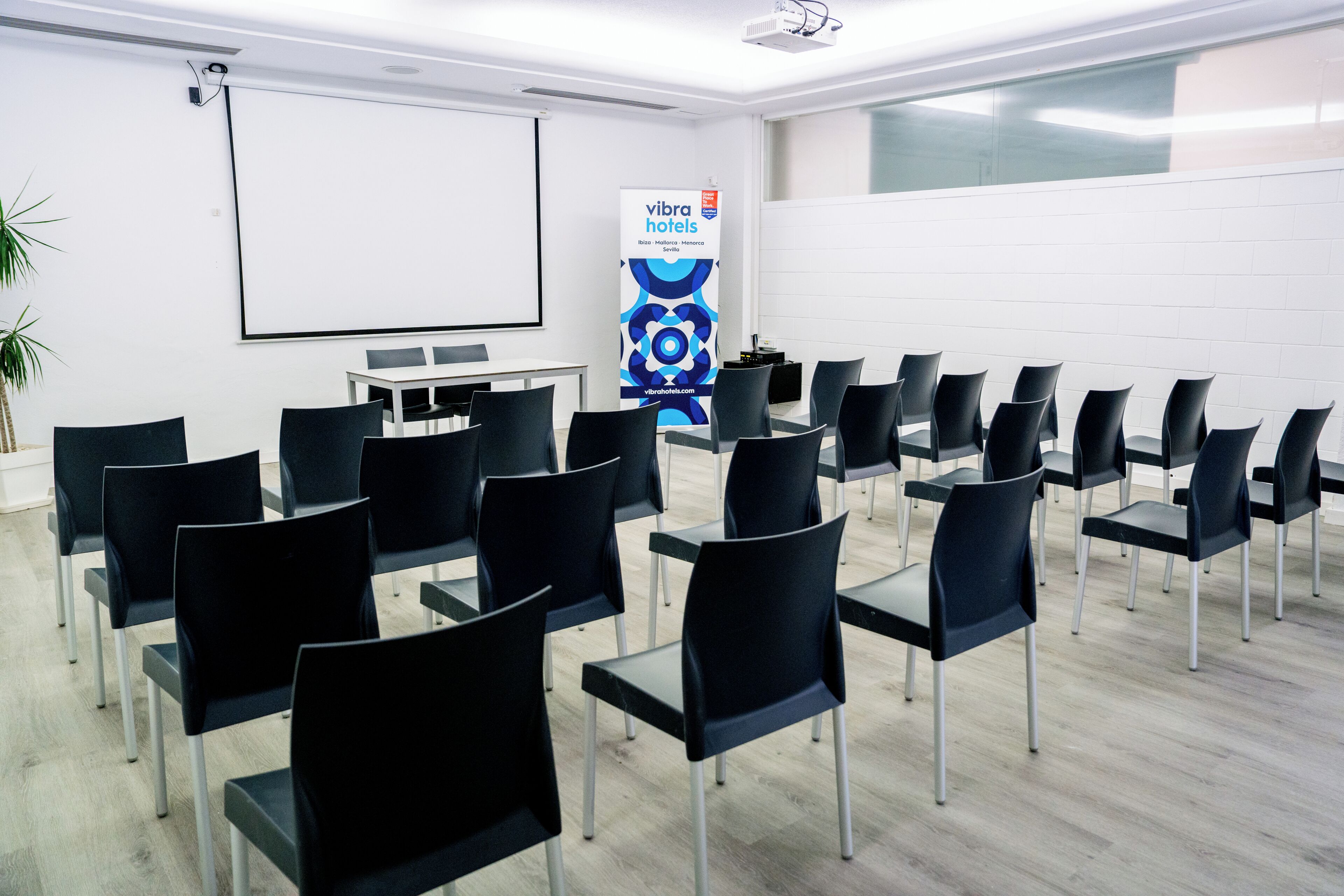 Un salón de conferencias con filas de sillas negras, una mesa blanca, una pantalla de proyector y un cartel para Vibration Hotels.