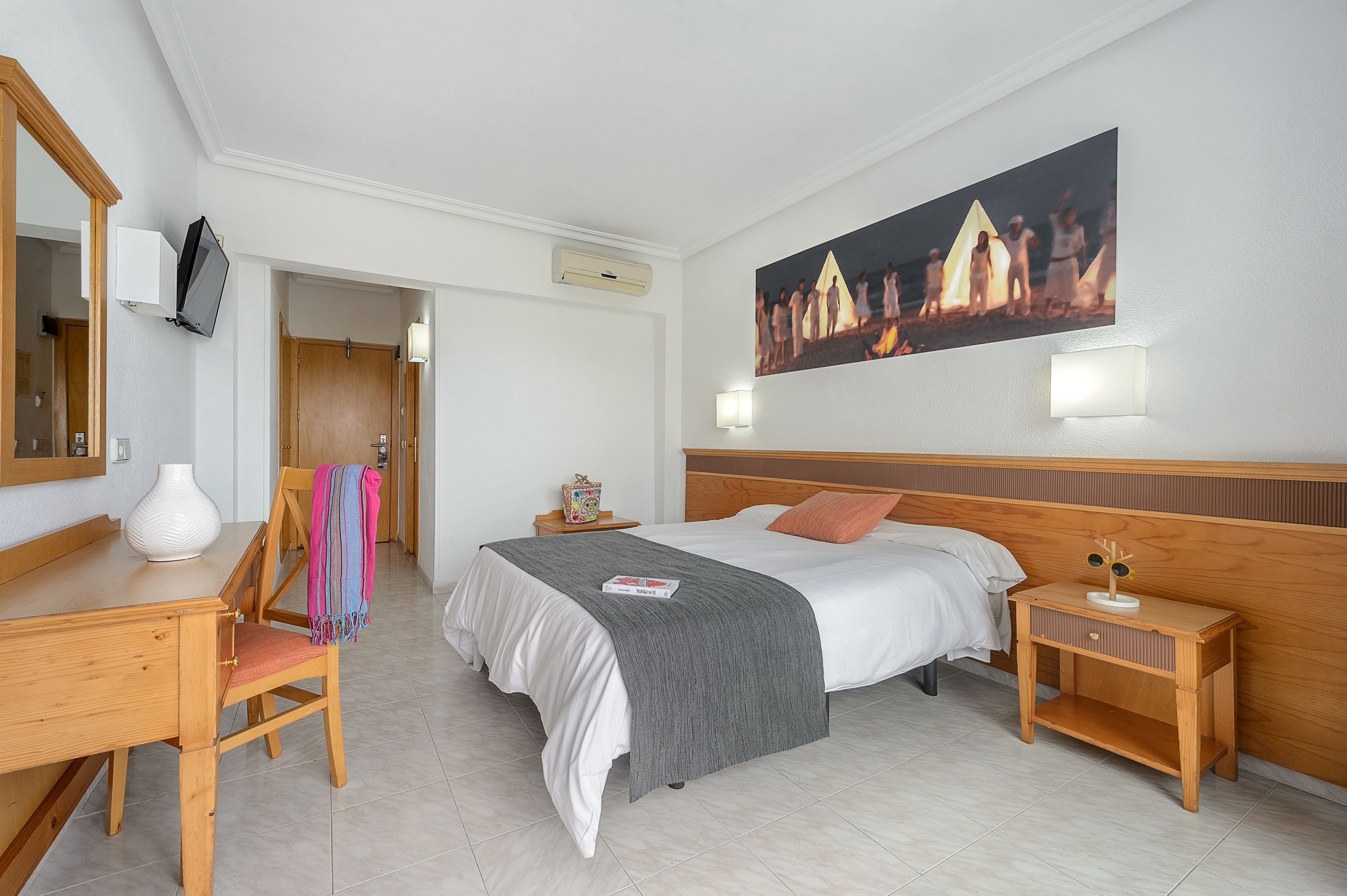 Habitación de hotel con cama, mesitas de noche, escritorio y una silla. Se observa un cuadro en la pared y un espejo sobre el escritorio.