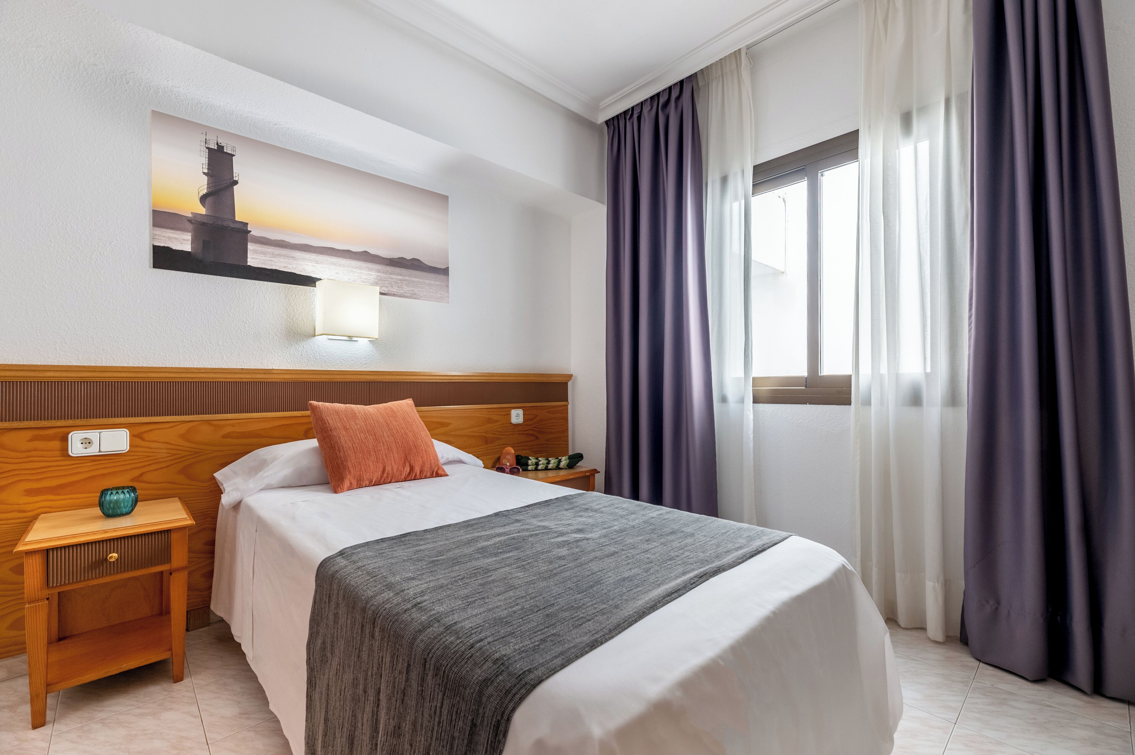Habitación de hotel ordenada con una cama, una mesita de noche, una lámpara y un cuadro de un faro en la pared.