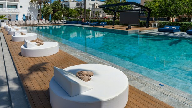 Un área junto a la piscina con sillones blancos circulares, una terraza de madera y una piscina azul cristalina.
