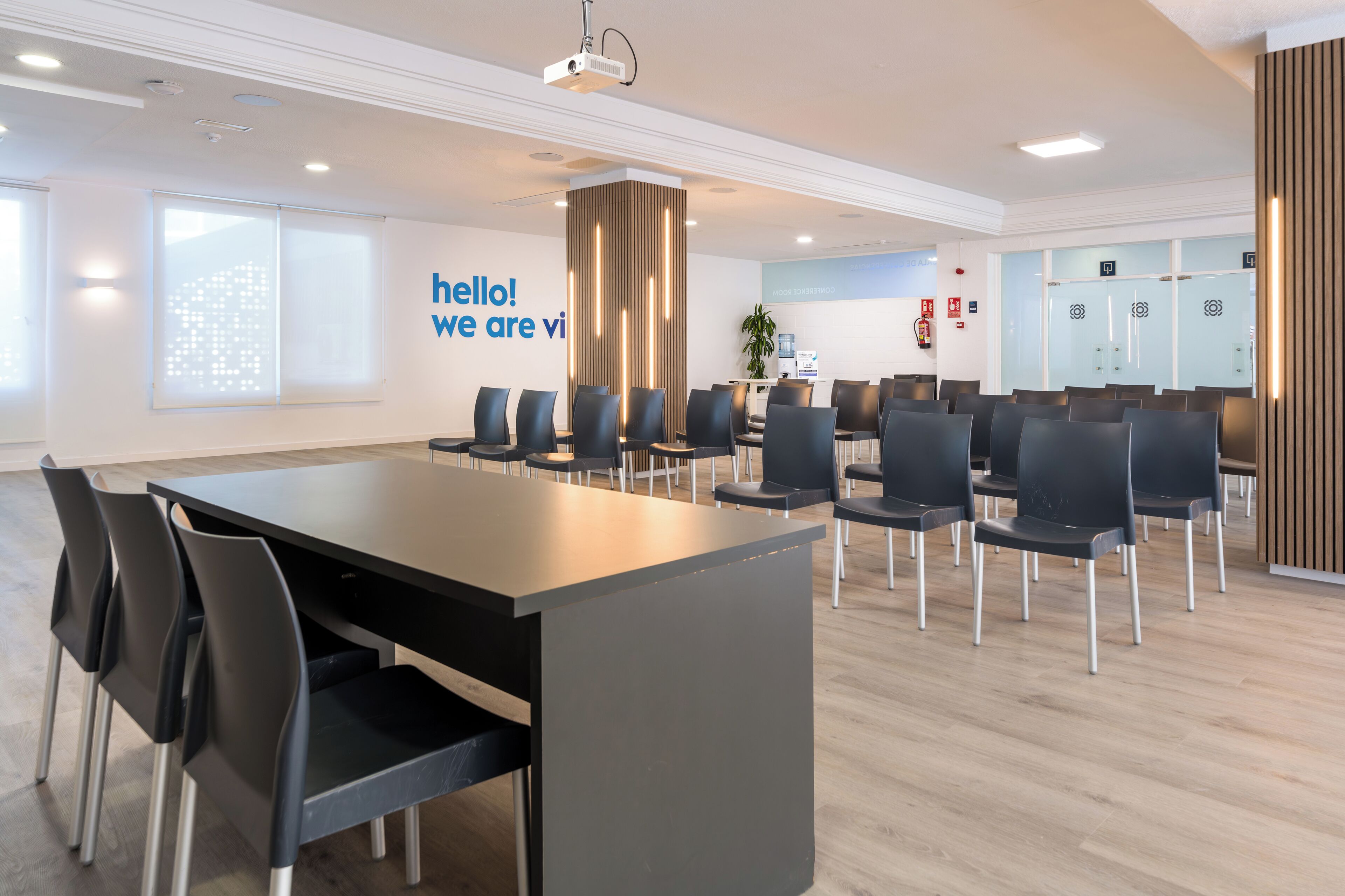 A modern conference room with a long table and chairs, a projector, and a 'hello! we are vi' sign on the wall.