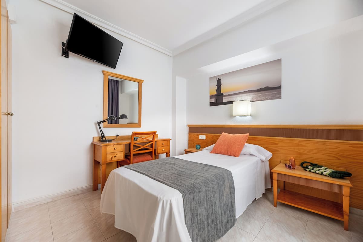 Una habitación de hotel con cama, escritorio con silla, televisor montado en la pared, un cuadro de un faro y una mesita de noche con lámpara y un bolso verde.