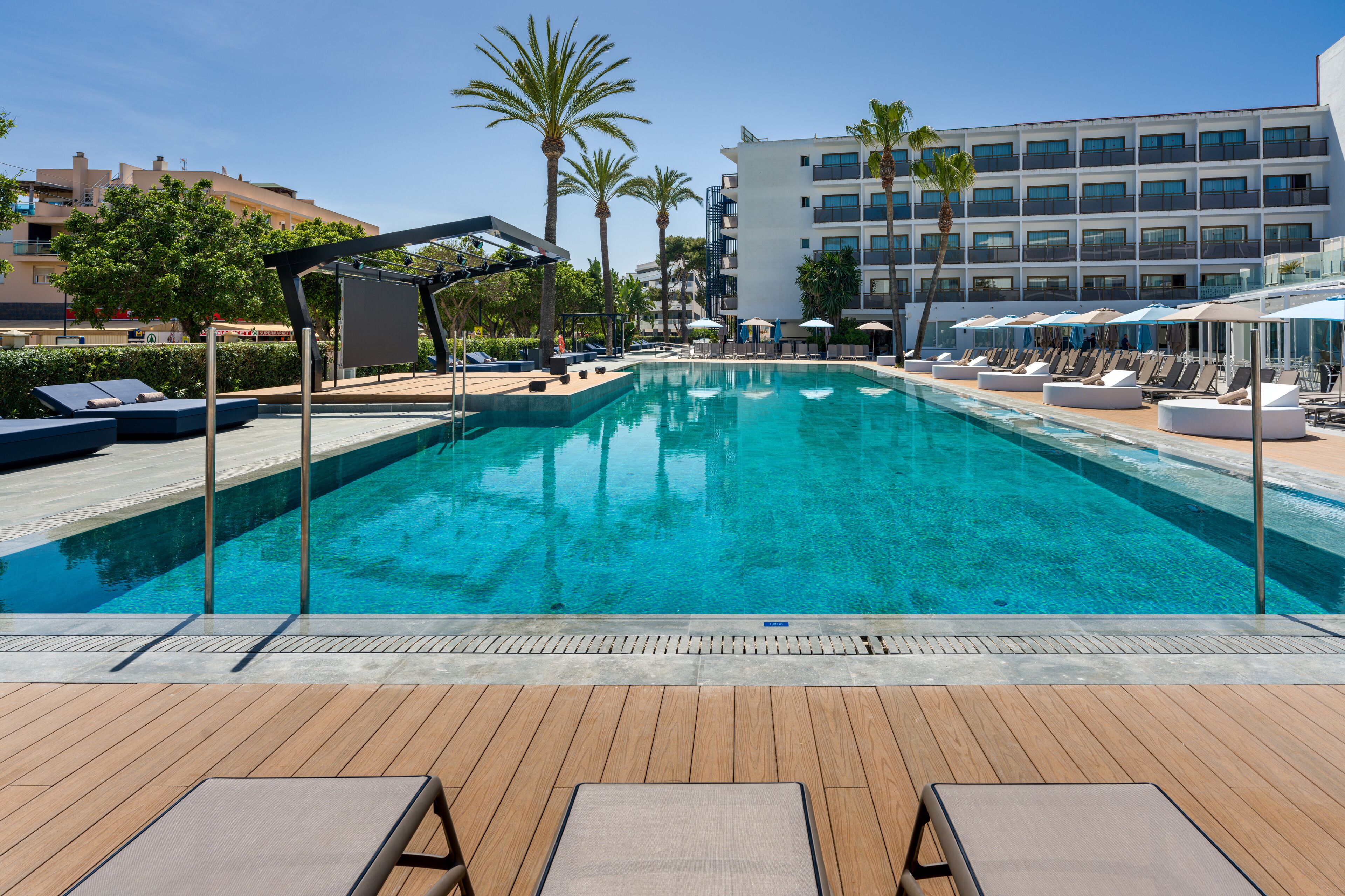 A swimming pool with lounge chairs and umbrellas, surrounded by palm trees and a hotel building.