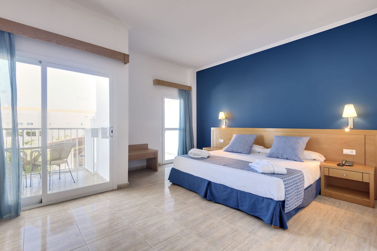 A bedroom with a bed, bedside tables, a nightstand, a window with curtains, and a balcony with chairs.