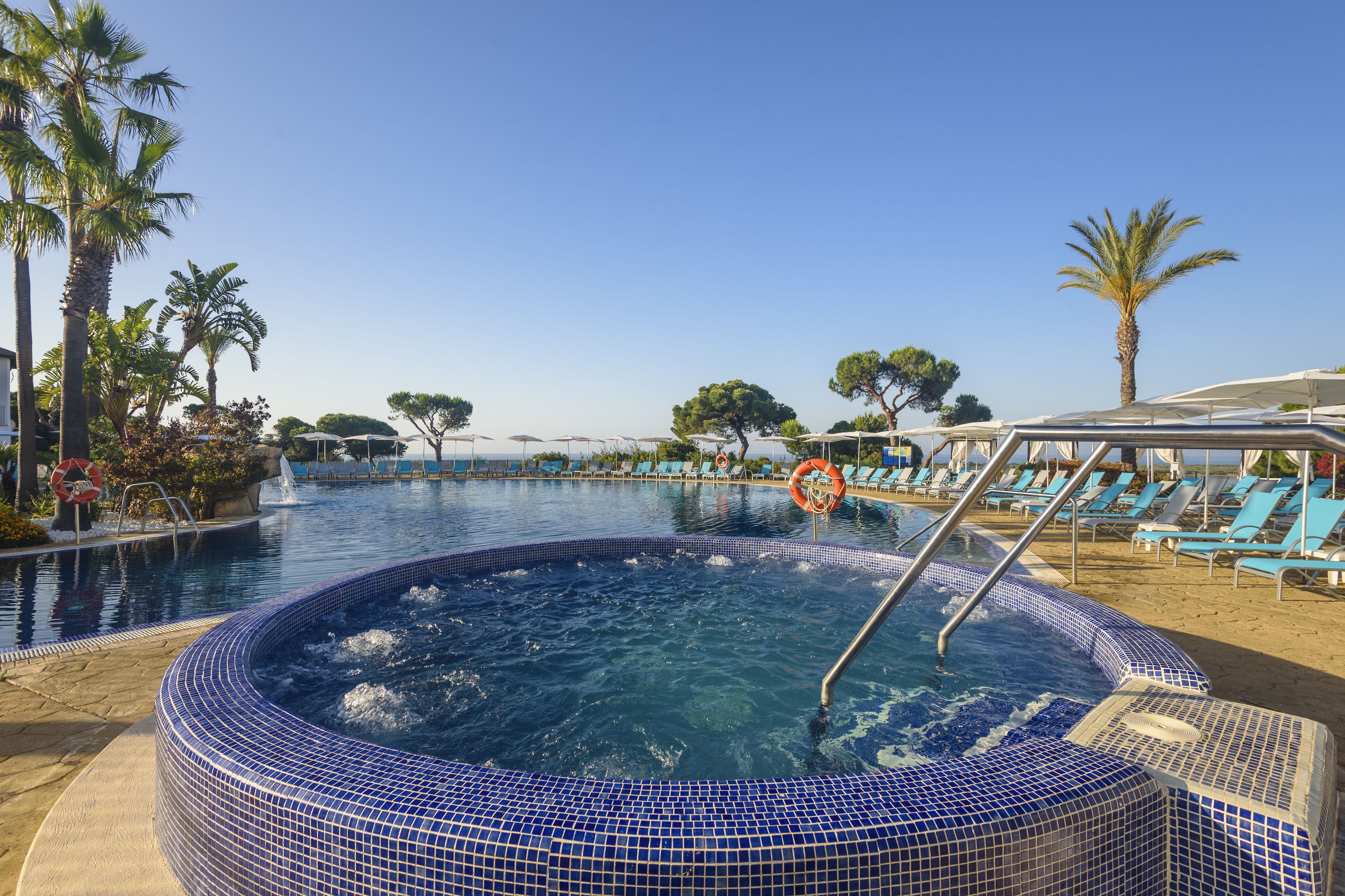 A swimming pool with a tiled hot tub, lounge chairs, palm trees, and a clear sky.
