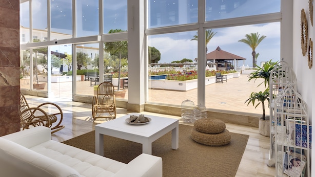 Amplia sala de estar con ventanas grandes, un sofá blanco, sillones de mimbre y una pequeña mesa.
