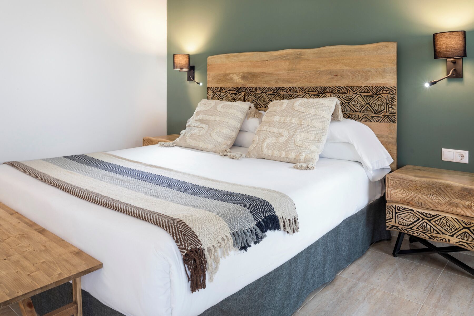 A neatly made bed with a patterned blanket, a wooden headboard, and two wall-mounted lamps.