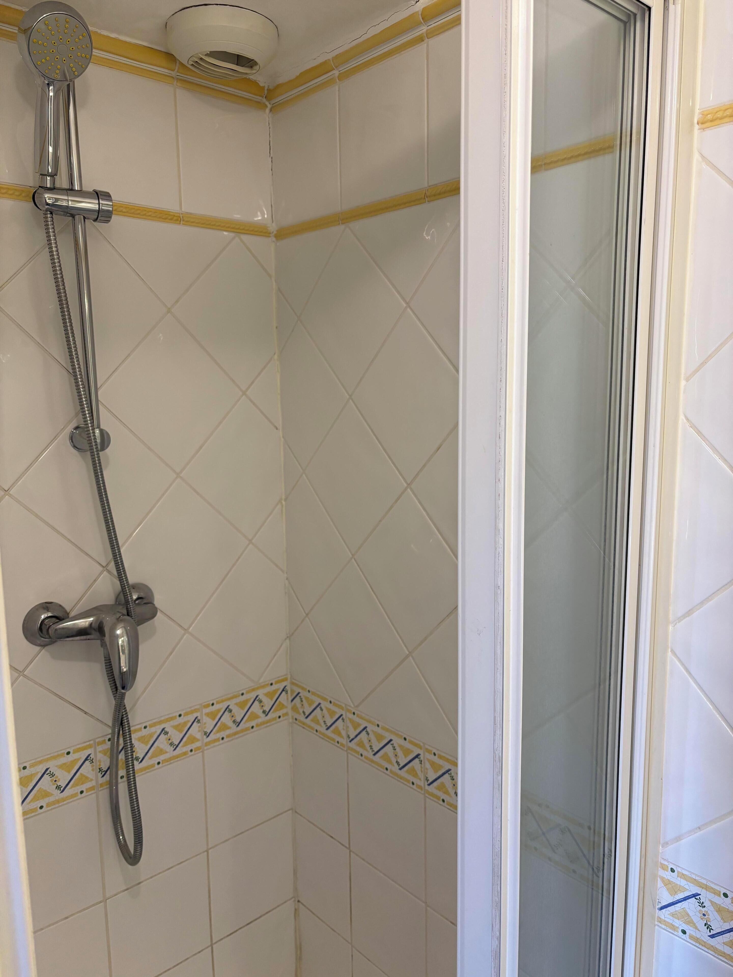 A shower with a handheld showerhead and a fixed showerhead, a corner with decorative tiles, and a glass door leading to a shower area.