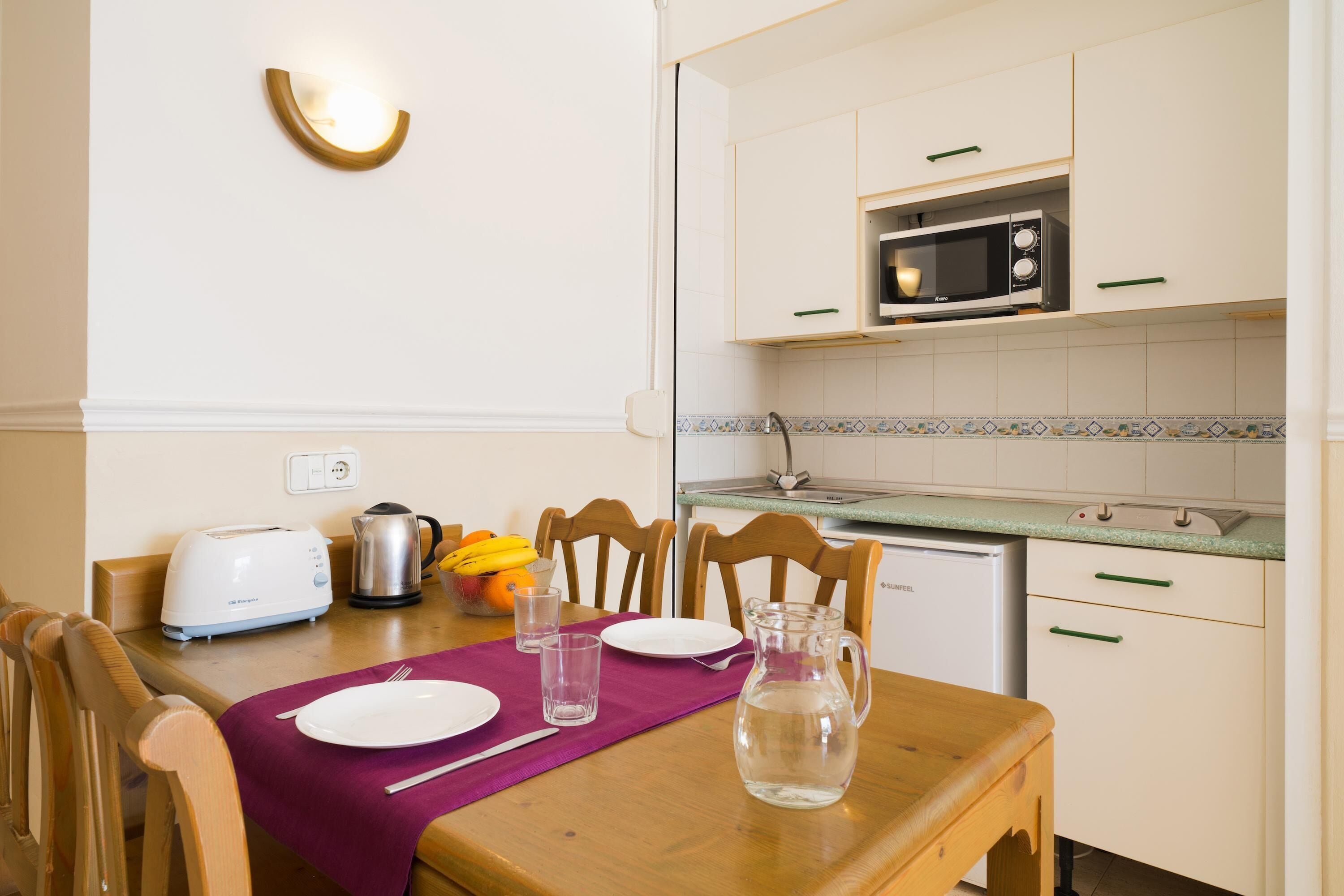 A clean, well-lit kitchen with white cabinets, a microwave, a toaster, and a dining table set with plates, glasses, and a pitcher of water.